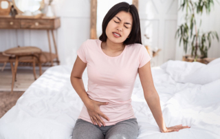 Woman sits on bed feeling pelvic pain and abdominal pain.