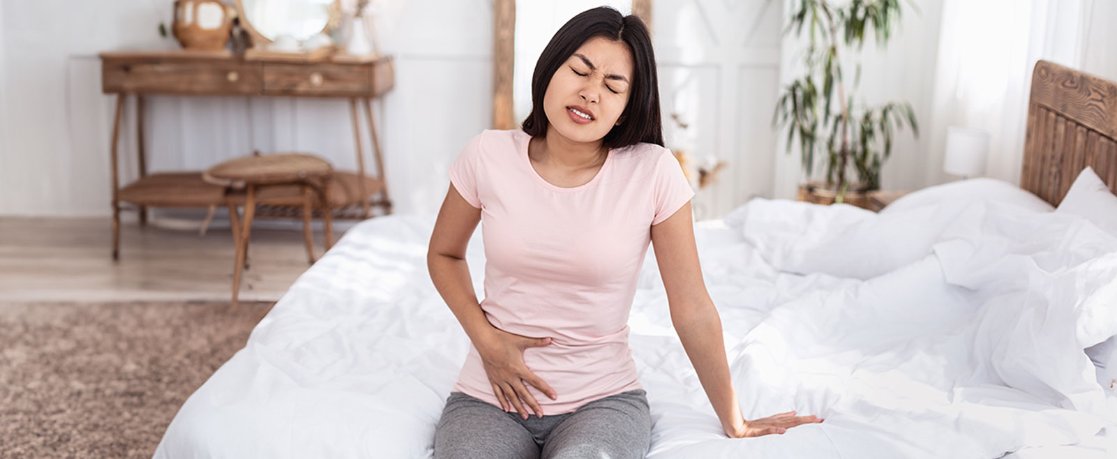 Woman sits on bed feeling pelvic pain and abdominal pain.